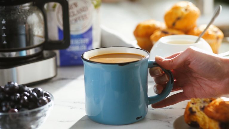 hand holding a fresh mug of coffee next to blueberry muffins and a pot